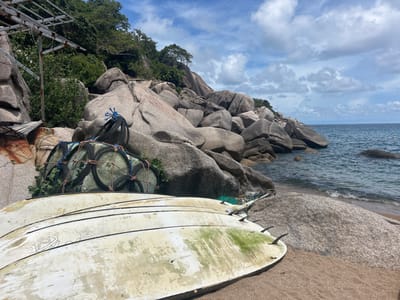 This image depicts a coastal scene in ตำบล เกาะเต่า, Thailand. In the immediate foreground, two weathered white surfboards lie upside down on the sandy beach; the upper board displays faint black graphics, and the lower one exhibits significant green algal growth, particularly around its three black fins. Behind these, a large, cylindrical fishing trap, fashioned from netting stretched over a framework of repurposed tires or rubber rings secured with blue rope, rests on the sand, partially intertwined with green foliage. To the far left, a dilapidated wooden structure with rusted corrugated metal panels is partially visible, topped by a rusty metal framework, possibly a boat hoist. The midground is dominated by an extensive formation of large, smooth grey boulders that descend towards the calm, dark blue ocean, where small waves gently break upon the shore. Lush green tropical vegetation grows on and between the boulders further inland. The sky above is partly cloudy, featuring scattered white and grey clouds. No individuals or animals are present in the scene.