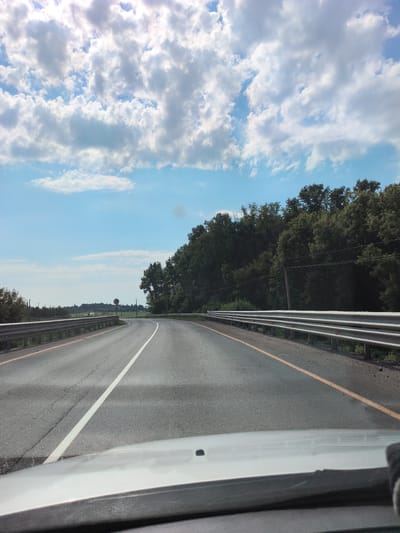 The image is a forward-facing view from inside a vehicle, likely a car, as it travels along a paved road. The road curves to the right and is marked with a white line on the left and a yellow line on the right. A metal guardrail runs along the right side of the road. On the left side of the road, there is a field with some vegetation and distant trees. The right side of the road is bordered by a dense line of trees. The sky above is blue with scattered white clouds. The overall scene suggests a rural or suburban environment. The image is not detailed enough to confirm the specific location within Polibino, Russia, but it depicts a typical road scene in such a setting. There are no people or specific actions visible in the image.