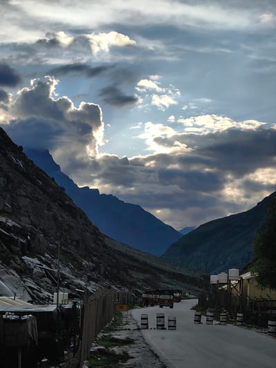 The image depicts a mountainous landscape with a road cutting through it. The sky is dramatic, with a mix of dark, heavy clouds and areas where sunlight breaks through, creating bright, ethereal rays. The mountains on either side of the road are steep and rocky, with some sparse vegetation visible.

The road itself appears to be a two-lane highway, with several vehicles visible. In the foreground, there are several white barrels with dark markings, possibly serving as road barriers or markers. A fence runs along the side of the road, and some small structures or shelters are situated behind it. Further down the road, a convoy of trucks is lined up.

A yellow sign with the letters "TCP" is visible on the right side of the road. In the distance, more mountains are visible, fading into a hazy blue. The overall impression is one of a remote, rugged location, possibly a mountain pass or a remote road in a high-altitude region. The lighting suggests it is either early morning or late afternoon, with the sun casting long shadows.

While the image does not explicitly show the city of Damphug, India, the environment is consistent with the terrain found in mountainous regions of northern India, such as Himachal Pradesh or Uttarakhand, where places like Damphug might be located. The presence of trucks and what appear to be checkpoints or barriers suggests this is a significant transportation route.