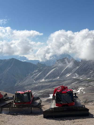This image depicts a bright, sunny day in the Austrian Alps, likely near the town of Wildermieming. In the foreground, two bright red snow groomers are parked on a gravelly terrain. These vehicles have large treads and a distinctive red cab with multiple lights. The machine on the right has a red mechanical arm extended.
Behind the snow groomers, a rugged mountainous landscape unfolds. The mountains are characterized by steep, rocky slopes and patches of snow, indicative of a high-altitude environment. Some of the peaks are partially obscured by wispy white clouds that are drifting across the bright blue sky. The overall scene suggests a location dedicated to winter sports or mountain maintenance, given the presence of the snow groomers.