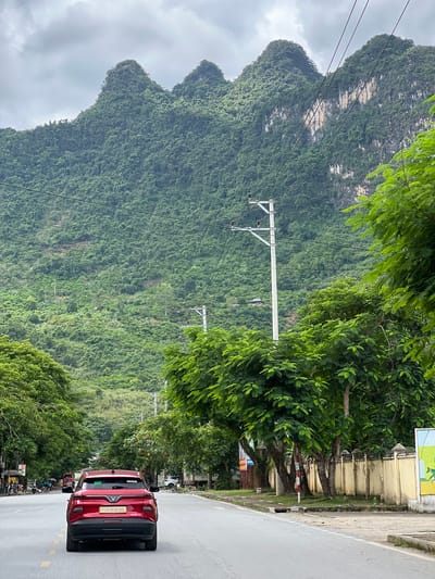 The image shows a road in the city of Hà Giang 2 Ward, Vietnam, with a red car driving away from the camera in the foreground. The road is lined with trees on both sides, and a large, lush green mountain range dominates the background. The sky is overcast. There are power lines and utility poles visible along the road. In the distance, there are some buildings and more vegetation on the mountainside. There are a few people and motorcycles on the side of the road in the far distance, near some market stalls. A yellow wall with decorative elements runs along the right side of the road. The overall scene depicts a scenic road in a mountainous region.