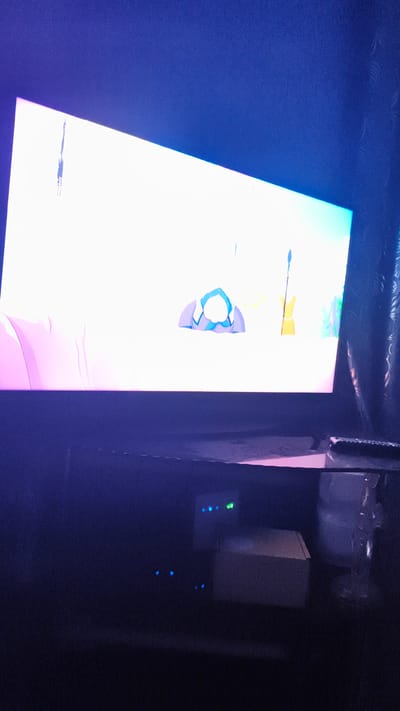 The image shows a television screen displaying animated content with a bright, abstract background and stylized figures. The screen is positioned above a dark entertainment unit. On the entertainment unit, there are several electronic devices with illuminated indicator lights, and a white rectangular object. A remote control is visible on the right side of the entertainment unit. The overall scene is dimly lit, with the primary light source being the television screen. The location is stated to be the Байқоныс ауылдық округі, Kazakhstan.