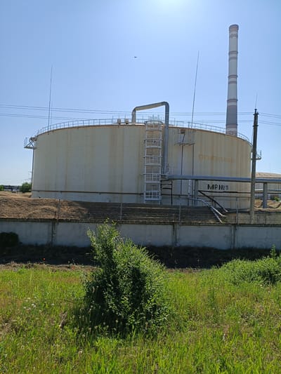 An industrial facility is depicted under a clear blue sky. In the foreground, lush green grass and a prominent bush create a natural element contrasting with the industrial structures. A concrete wall with a barbed-wire fence runs across the mid-ground, separating the natural vegetation from the industrial area.

Dominating the scene is a large, cylindrical, light-colored tank with a railing around its perimeter. A metal ladder ascends one side of the tank, and pipes are connected to its surface. To the right of the tank, a tall, striped smokestack rises into the sky. The smokestack is segmented with white and reddish-brown bands.

Further industrial elements, including more pipes and structures, are visible in the background. The overall scene suggests an operational industrial site, likely a power plant or processing facility, located in Dnestrovsk, Moldova. The presence of power lines and the sheer scale of the infrastructure indicate a significant industrial presence. The lighting suggests it is daytime, with the sun high in the sky.