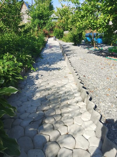 The image shows a garden path in Iskitim, Russia. The path is constructed with irregularly shaped concrete pavers, bordered by a wavy concrete edge on one side and gravel on the other. Lush green foliage, including hosta plants and trees, line the path. In the background, a wooden structure, possibly a house or shed, is visible. The scene is illuminated by bright sunlight, casting shadows on the path and plants. There are no people or animals present in the image.
