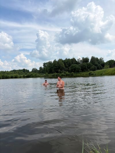 Two men are swimming in a wide, calm lake surrounded by dense green trees and vegetation under a cloudy sky. The man on the left is further from the viewer, and appears to be looking away from the camera. The man on the right is closer to the viewer, and is facing towards the left. His body is mostly submerged in the water. The water is dark and reflects the sky and trees. The scene is peaceful and natural, suggesting a remote or rural setting.