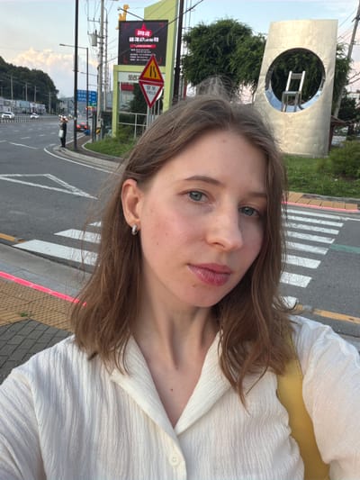 In Pocheon-si, South Korea, a young woman with light brown hair and blue eyes is taking a selfie in front of a pedestrian crossing. She is wearing a white collared shirt and a yellow bag is visible over her shoulder. Behind her, a busy street with cars and traffic lights stretches into the distance. A large, abstract metal sculpture with a circular opening and a chair visible inside stands to the right of the woman. A triangular yield sign with Korean text is also present in the background, along with a billboard featuring Korean writing. The sky is a pale blue with scattered clouds, suggesting it is late afternoon or early evening.