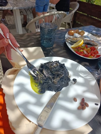 An overhead, close-up shot shows a white plate of black squid ink risotto with a bright green sauce on the left, being forked by a woman with white-tipped nails and light skin. To the right of the risotto, a knife rests on the plate, next to a small pile of nuts. In the background, a white plate holds a dish of fried fish, a lemon wedge, and chopped red and green peppers. A blue glass sits behind the plate of risotto, and a shadowed table fills the foreground. In the background, two people wearing denim shorts and white chairs are visible, suggesting an outdoor dining setting. The scene appears to be a restaurant in Sines, Portugal, possibly along the coast, given the presence of seafood.