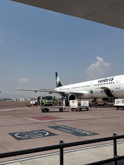 The scene depicts an active airport apron at Guadalajara International Airport (GDL) in Zapote del Valle, Mexico, under clear daylight conditions.
A white Volaris passenger aircraft, identified by "Volaris" text on its fuselage and a distinctive colorful, pixelated pattern on its vertical stabilizer, is parked on the tarmac. The registration "EI-MCO" is partially visible near the rear of the fuselage. Its forward cargo door is open, and a baggage belt loader is actively engaged, suggesting cargo loading or unloading operations are underway.
Essential ground support equipment surrounds the aircraft. A green utility truck is positioned near the aircraft's right engine. Several white baggage carts/dollies are present, one prominently displaying "GDL-CX-134" and another in the background marked "TAXIS-GP-GDL." At least two ground crew personnel are observed near the green truck and the engine area. In the background to the left, multiple portable passenger stairs are staged.
The ground consists of an asphalt tarmac with various painted markings, including a circular pattern in the immediate foreground and faint text on a dark rectangle. A black metal railing runs across the bottom of the frame, indicating the vantage point is from an elevated structure, likely part of a terminal. Distant mountains are visible on the horizon under a predominantly clear blue sky with minimal cloud cover.