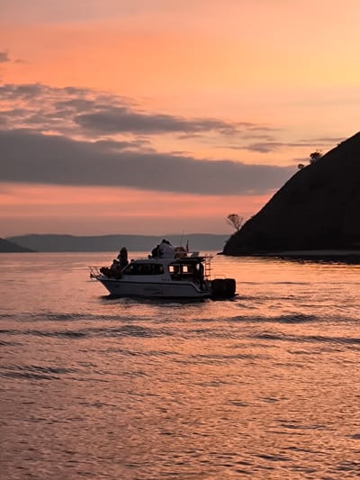 A boat carrying several people sails on the water during sunset. The sky is a gradient of orange and pink, with scattered clouds. A dark, tree-covered hill juts out from the right side of the frame, meeting the water's edge. In the distance, hazy mountains are visible on the horizon. The water is calm, with gentle ripples reflecting the colors of the sky. The boat is white with a cabin and appears to be a tour boat. Some passengers are standing on the deck. The scene evokes a sense of peacefulness and the beauty of a tropical sunset. The time of day is clearly sunset due to the colors in the sky and the lighting. The location appears to be a coastal area with islands or a mainland visible in the distance, consistent with Rinca Village, Indonesia. Visible text on the boat reads "COLUMBIA".