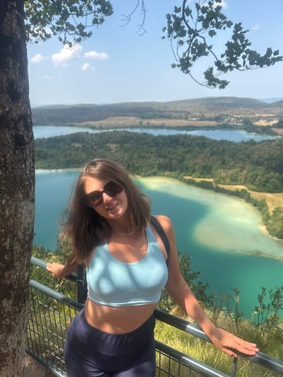 A woman with brown hair and sunglasses is standing on a railing next to a tree. She is wearing a light blue sports bra and dark blue leggings. Behind her, there are several bodies of water and a forested landscape under a clear blue sky. The scene appears to be a scenic viewpoint overlooking lakes and hills in the commune of La Chaux-du-Dombief, France.