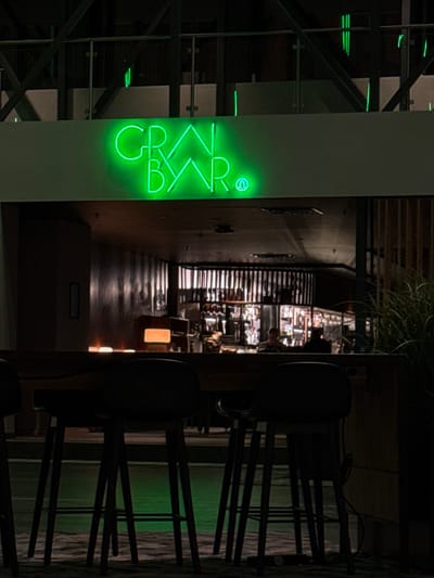 A neon green sign reading "CR BYR" vertically illuminates the exterior of a building. Beyond, a narrow corridor stretches into the distance, featuring a bar or restaurant with seating and shelving. To the right, chair backs are visible, along with an outdoor area with vegetation and fencing. The photograph appears to be taken at night.