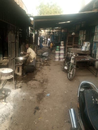 The image displays an unpaved market alleyway in Kofar Ruwa, Nigeria, under natural daylight. The ground is uneven dirt with visible damp spots and scattered refuse. On the left side, a person with short hair, wearing a light-colored long-sleeved top and dark trousers, is actively washing items in a large, round metal basin positioned on a low wooden stool. A dark pot rests directly on the ground beneath the basin. To the immediate left of this activity, a makeshift stand constructed from a metal wheel rim is visible.
The alley is bordered on both sides by open-fronted market stalls or shops, characterized by metal grates and covered by corrugated metal awnings. On the right side of the alley, a tall stack of commercially labeled white and green paint buckets is prominently displayed. A light-colored, older model motorcycle with a single headlight is parked facing left in the mid-ground on the right. Further into the foreground on the right, the rear portion of a dark motorcycle, including its seat and a chrome exhaust pipe, is partially visible, suggesting the camera's position.
Beyond the immediate activity, several individuals are present further down the alley, including one person in a dark blue garment and another near a small bicycle. The market structures extend into the distance, where trees and brighter light indicate an opening or less dense area beyond the covered stalls.