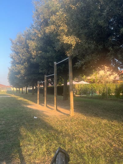 The image depicts an outdoor scene with rows of trees casting shadows on a grassy area. In the foreground, there are several horizontal bars supported by wooden posts, resembling an outdoor fitness equipment setup. The ground is covered with grass, and there is a small piece of white debris on the ground. In the background, there are more trees, a hedge, a fence, and some buildings with umbrellas visible. The lighting suggests it is either early morning or late afternoon, with the sun casting long shadows. The location is identified as Sant'Eufemia, Italy.