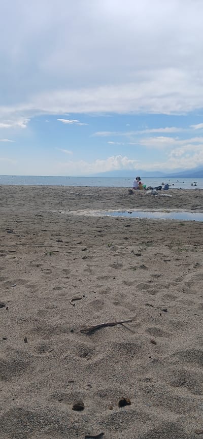 In Priozernoe, Kyrgyzstan, on a sandy beach, two individuals sit near the water's edge. One person, in a white shirt, is hunched over, possibly attending to something on the sand. Beside them, another person, wearing a yellow shirt, is also seated. The beach is sparsely populated with a few other figures visible in the water, swimming or wading. The water is calm and reflects the light blue sky, which is dotted with white clouds. In the distance, a hazy mountain range lines the horizon. The sand in the foreground is textured, with small pebbles and a few scattered pieces of driftwood. A small puddle of water is visible on the sand, reflecting the sky.