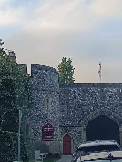 The image depicts the exterior of Arundel Castle, located in Arundel, United Kingdom. The castle is constructed of stone with a grey, textured facade. A large, arched wooden entrance is visible on the right side of the image, leading into the castle. Adjacent to this entrance, to the left, is a smaller, red-painted door. A sign on the stone wall to the left of the red door displays information about Arundel Castle & Gardens, including opening dates and times, a website address, and a telephone number.

A flag, white with a red cross, flies from a flagpole atop the castle wall. Trees with green foliage are visible on the left and right sides of the castle, partially obscuring the structure. In the foreground, the rear of a white vehicle is parked, partially obscuring the lower portion of the castle entrance. A wooden bench is situated on the left side of the image, in front of the castle wall and surrounded by greenery. The sky above is overcast with light-colored clouds.