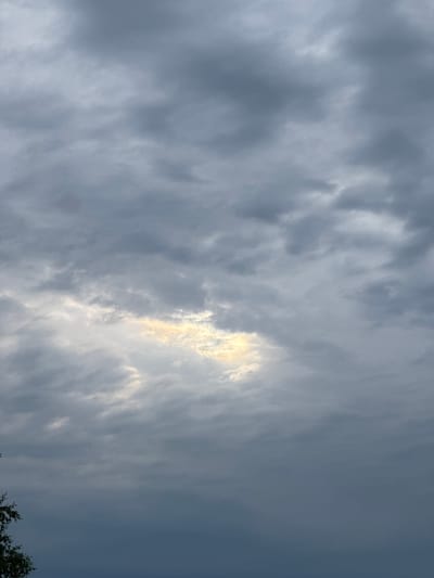 The image depicts a cloudy sky with a patch of sunlight breaking through the overcast. The clouds are predominantly gray and appear to be layered. The lighting suggests it is either early morning or late afternoon. The bottom left corner of the image shows the top portion of a tree with green leaves. The scene appears to be an outdoor environment in Володарский ауылдық округі, Kazakhstan, characterized by a natural landscape. There are no people or specific urban structures visible.