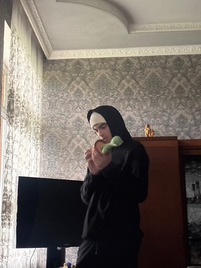 The image depicts an indoor residential scene in Durgena, Georgia. A single individual, appearing to be a young male, stands centered in the frame, facing slightly right. He wears a white beanie under a black hooded sweatshirt and black sweatpants. The individual holds a light brown, circular object, possibly a bun or small cake, in both hands, accompanied by a small, light green plush toy positioned atop the circular object.

To the left, a window covered by a translucent white curtain with intricate patterns allows natural light into the room. A large, dark-screen television is visible in the lower-left background. To the right, a tall, dark wooden cabinet with glass doors stands, featuring a golden-colored decorative item on its top surface. The walls are covered with wallpaper displaying a repeating grey and beige damask pattern. The ceiling is white with decorative crown molding along the perimeter.