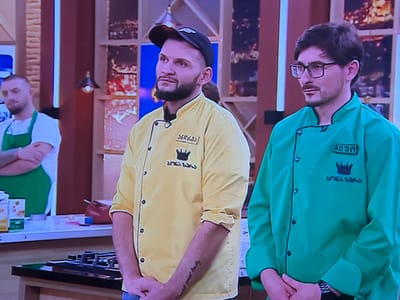 The image depicts three males in a brightly lit studio setting, presented as a kitchen environment, located in the city of Colombes, France.
In the foreground, two men stand side-by-side, facing generally forward. The man on the left wears a yellow chef's jacket with black buttons, a black collar, and a black baseball cap. He has a dark beard and mustache and is looking slightly to his right. His jacket features Georgian script, including "SERGI" (სერგი) above a crown logo, and "MASTERCHEF" (მასტერშეფი) below it. His hands are clasped in front of him.
To his right, the second man wears a green chef's jacket with black buttons and collar. He has dark hair, glasses, and a neatly trimmed mustache and beard. He looks directly ahead, and his jacket also displays Georgian script, with "ADAM" (ადამ) above a crown logo and "MASTERCHEF" (მასტერშეფი) below it. His hands are also clasped in front of him.
Partially visible in the background on the left is a third man with a beard, wearing a white t-shirt and a green apron, standing with his arms crossed, observing the scene.
The immediate foreground features a black countertop with what appears to be a built-in stovetop or cooking range. Further back on the left, a counter holds various kitchen items, including a white carton and a small red bowl. The background is a stylized studio set, featuring illuminated shelving units with segmented warm lights, some resembling city lights, and vertical light strips, contributing to a modern television production aesthetic.