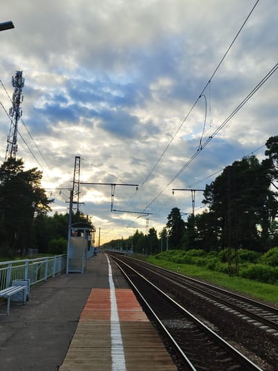 This image depicts a train station platform with an adjacent railway track under a cloudy sky. The platform is made of concrete and wooden planks, with a white and orange marking indicating a safety line. A metal fence and a small shelter with a bench are visible on the platform. Overhead, train power lines are strung between poles.
To the left of the tracks, a dense line of green trees and foliage forms the backdrop, with a tall cell tower structure visible in the lower left corner. The railway tracks themselves are set on ballast.
The scene appears to be set in the late afternoon or early evening, with the sun casting a warm glow through breaks in the clouds. The weather is overcast with scattered sunlight. Two figures are visible in the distance on the platform, suggesting the station is operational, though it doesn't appear crowded. The overall mood is tranquil and somewhat dramatic due to the contrast of light and shadow in the sky. The location is identified as Tsibino, Russia. No discernible text is present in the image.