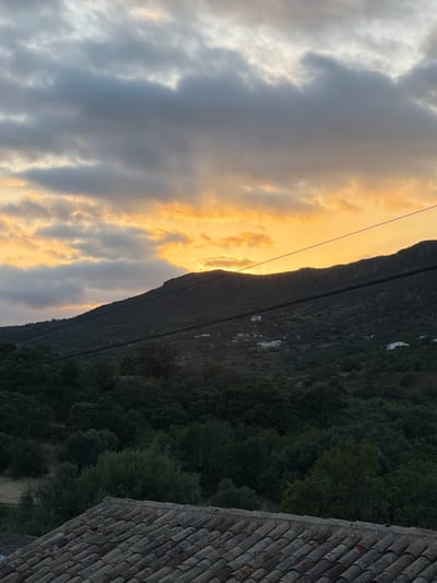 The image depicts a sunset over a hilly, wooded landscape in the city of Loulé, Portugal. In the foreground, a section of a tiled rooftop is visible. Beyond the rooftop, a dense forest covers the lower slopes of a mountain. Several white buildings are scattered across the mountainside. The sky above is filled with dramatic clouds, tinged with the orange and yellow hues of the setting sun, which is partially obscured by the mountain peak. Power lines stretch across the scene, adding a man-made element to the natural landscape. The overall impression is one of a rural or semi-rural setting at dusk.