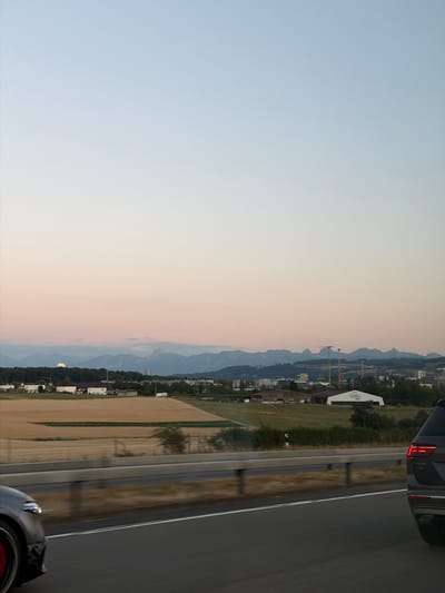 The image captures a panoramic view of a landscape, likely from a moving vehicle on a highway. The scene is divided into a vast, clear sky transitioning from a pale blue at the top to a soft pink and orange near the horizon, suggesting either sunrise or sunset.
Below the sky, a town or village is visible nestled against a backdrop of rolling hills and distant mountains. Some buildings appear to be residential, while others might be commercial or industrial. Construction cranes are noticeable in the mid-ground, indicating ongoing development. A prominent white building with the word "TIVOLI" printed vertically on its side is clearly visible.
In the foreground, a paved road, presumably a highway, runs alongside a field of dry, yellowish crops. The reflection of trees and the sky on the wet road surface suggests recent rain or morning dew. The rear of a dark-colored car is visible in the bottom right corner of the image, with its brake lights illuminated, confirming the perspective from a vehicle.
The overall mood is tranquil and serene, amplified by the soft light and expansive sky. The clear visibility and the presence of distant mountains place the location in a scenic, possibly alpine, region. The time of day is either dawn or dusk. The visible text "TIVOLI" on a building is a notable detail. The scene appears to be set in Ittigen, Switzerland.