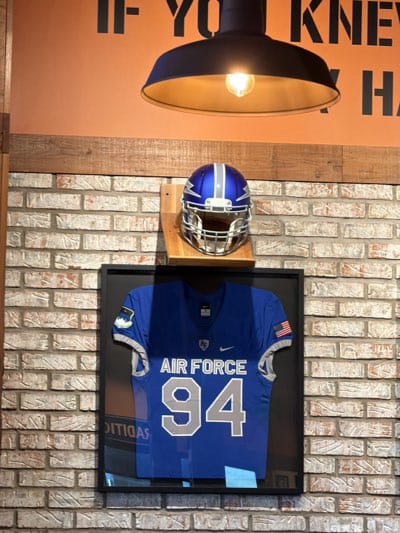 A framed Air Force jersey with the number 94 hangs on a brick wall, with an Air Force football helmet displayed on a small wooden shelf above it. A black industrial-style light fixture illuminates the scene from above, casting a warm glow. The wall is constructed of tan and gray bricks and is located in the city of Rochester, United States.