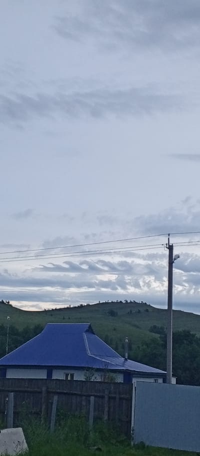 In Khusainovo, Russia, an overcast sky looms above a rural landscape. A blue-roofed house with white siding is partially visible behind a wooden fence, its roof featuring a unique, multi-pitched design. A utility pole with a light fixture stands to the right of the house. In the background, rolling green hills dotted with sparse trees stretch towards the horizon, their contours softened by the diffused light. The overall scene suggests a quiet, possibly remote, dwelling.