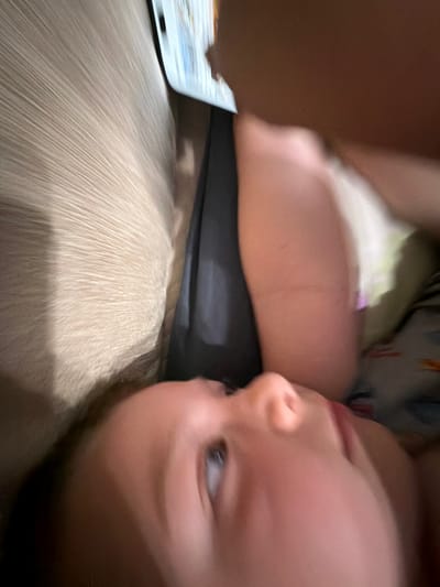 The image shows a close-up, slightly blurry view of a child's face, looking upwards. The child has light-colored skin and a visible eye. Part of the child's arm is also visible, appearing smooth and pinkish. Behind the child, a textured surface, possibly a fabric or upholstery, is visible, with faint lines running horizontally. To the right of the child, a dark fabric is present, and further to the right, another person's arm is visible, holding a tablet device. The tablet displays an image that is difficult to discern due to the blurriness. The overall scene appears to be indoors, possibly in a living room or bedroom setting. There is no information available to determine the location of Zmeitsa, Bulgaria.