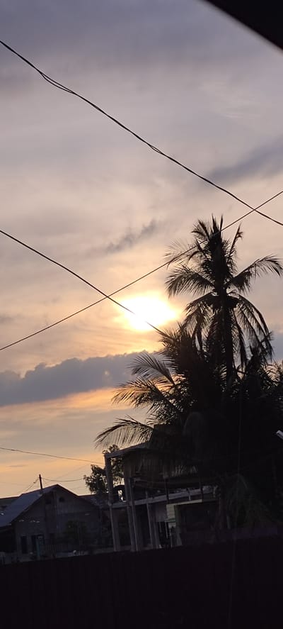 The image depicts a sunset scene in Dayahlhoksukon Barat, Indonesia. The sky is filled with soft, diffused light, with hues of orange and pale yellow indicating the setting sun, which is partially obscured by clouds. Silhouetted against the sky are the fronds of a coconut palm tree, characteristic of tropical locations. In the foreground, power lines crisscross the frame, suggesting an urban or semi-urban environment. Below the sky and palm tree, the silhouettes of several buildings are visible. One building appears to be a house with a sloped roof, while another is a partially constructed structure with concrete pillars and beams. The overall mood is tranquil and serene, with the soft lighting of the sunset contributing to a peaceful atmosphere. The time of day is clearly evening, as suggested by the sunset. No people are visible in the image. There is no visible text.