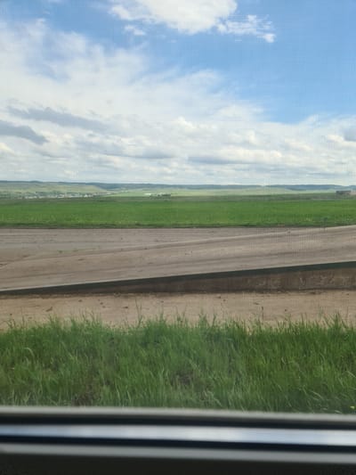 This media file is a vertical landscape image captured from a window, showing a view of the countryside in Petrovka, Russia. The foreground features a concrete path running diagonally across the frame. To the left of the path is a vibrant green field, likely farmland, stretching towards the horizon. Beyond the field, a small village with scattered houses can be seen nestled among rolling hills. Further in the distance, more undulating terrain is visible, all under a bright blue sky filled with fluffy white clouds. To the right of the concrete path, a strip of lush green grass grows along the edge of the path. The right edge of the image is partially obscured by what appears to be a window frame, showing vertical black and white blinds. The lighting suggests it is daytime, likely mid-morning or afternoon, with clear and pleasant weather. The scene depicts a peaceful rural setting. There is no visible text in the image.