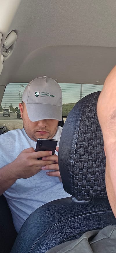 The image depicts a man wearing a light grey baseball cap with the logo "Air Products SAFETY ACADEMY Hosted by AUT" in green and black. He is looking down at his black smartphone, holding it with both hands. He is wearing a light grey t-shirt. The scene appears to be inside a vehicle, with the headrest of a black leather seat visible in the foreground on the right. The background shows a window with a view of a road with a few cars and some vegetation, suggesting an outdoor environment likely on the outskirts of a city. The man is seated in what appears to be the passenger seat of the vehicle.