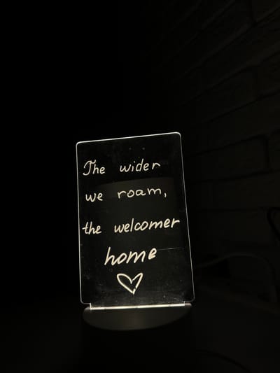 An illuminated square sign with handwritten white text and a heart symbol sits on a dark surface. The text reads: "The wider we roam, the welcomer home." The sign is positioned on a slightly reflective, dark surface, possibly a tabletop, against a dark background with a faint texture suggestive of wooden planks. The sign appears to be made of clear material, possibly acrylic, with LED lighting emanating from its base, casting a subtle glow upwards and around the edges. No people are present in the image. The overall scene is dimly lit, emphasizing the glowing sign. The location of Podilsk, Ukraine, is not evident from the visual information in the image.