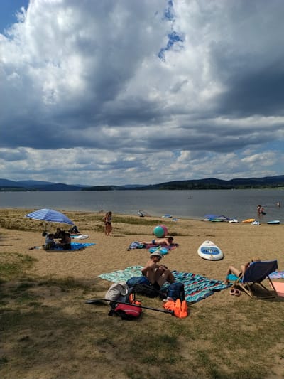 The image depicts a sunny day at a beach with a large body of water and hills in the background, suggesting a location like the Radslav Reservoir in the Czech Republic. Several people are present on the sandy beach, some of whom are sunbathing on towels and blankets. An umbrella provides shade for a small group of people. Various water sports equipment, including paddleboards and a large inflatable ball, are scattered on the sand. The sky is filled with dramatic clouds, indicating a dynamic weather system. The overall atmosphere is one of leisure and outdoor recreation.