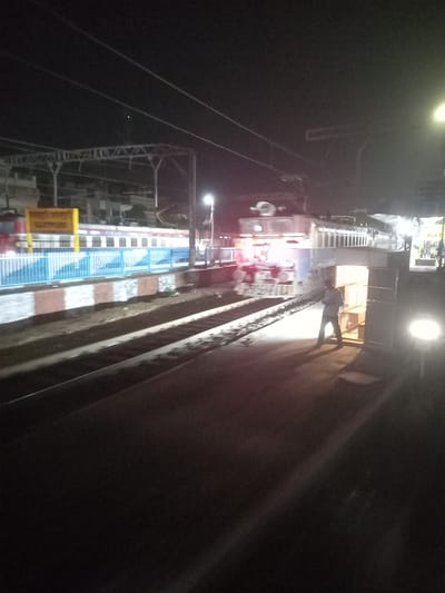 The image shows a train station platform at night. A train is stopped at the platform, its carriages visible with a light blue and white pattern. A yellow sign is on the platform, possibly indicating the station name. A person walks along the platform, next to the train tracks. Overhead, there is a covered walkway and overhead lighting, and electrical lines. The location is Igatpuri, India.