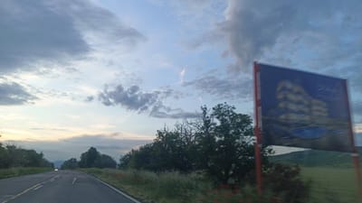 A slightly blurred, daytime photograph shows a paved road in Sadievo, Bulgaria. The road stretches towards the horizon, where a vehicle with headlights on is visible. The sky is a mixture of blue and gray clouds, indicating either early morning or late afternoon.

To the right of the road, vegetation is visible, including trees and grassy areas with red flowers. A large, blurred sign dominates the right side of the frame; its details are unclear but seem to depict a building and a body of water. Behind the vegetation and sign is a field, and a small hill in the distance.