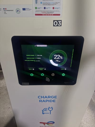 An electric vehicle charging station shows a display indicating 22% charge completion and 79 kW power. The charging details on the screen show an energy charged value of 0.2616 kWh and a charging time of 22 seconds, with an estimated time to reach 100% charge at 1 hour and 19 minutes. The station, labeled "CHARGE RAPIDE" (Fast Charge), features the TotalEnergies logo. A contact number is displayed for assistance, and there are instructions for operating the station. The charger's identification number is 03.