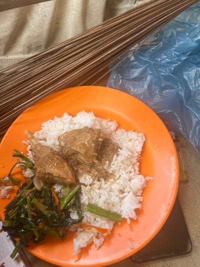 The image shows a plate of food on an orange plate. The food consists of white rice, some meat in a brown sauce, and green vegetables. In the background, there is a pile of dry brown sticks, a crumpled blue plastic bag, and a beige fabric. A black smartphone is visible beneath the plate. The scene appears to be set outdoors or in an open-air location. The overall impression is of a simple, rustic meal. The location is stated to be Matangwakeuh, Indonesia.