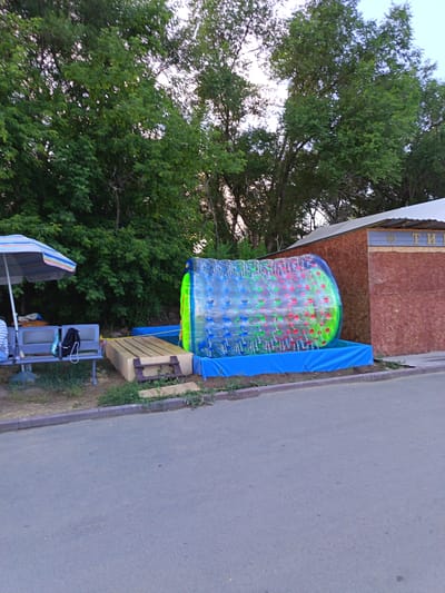 This image depicts an outdoor scene in Yarovoye, Russia, possibly at an amusement area or park. In the foreground, a paved surface transitions to a grassy area with a raised curb. To the left, a striped umbrella shades a blue bench, on which a backpack and a water bottle are placed. Behind the bench, a large, transparent inflatable sphere filled with red and green plastic balls is partially submerged in a blue inflatable pool. The sphere is positioned in front of a wooden structure with a corrugated metal roof. Trees form a dense green backdrop. The overall scene suggests a recreational setting, likely operational during daylight hours.