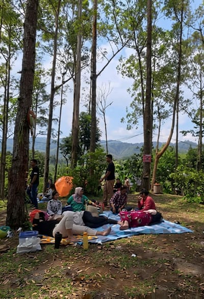 The image depicts a group of people enjoying a picnic in a forested area. The setting appears to be Laugumba, Indonesia, with lush green trees and rolling hills visible in the background. Several individuals are seated or lying on blankets spread out on the ground, engaged in various activities such as relaxing and conversing. An orange tent is pitched nearby, suggesting a camping or day-trip excursion. The overall atmosphere is one of leisure and enjoyment amidst nature.