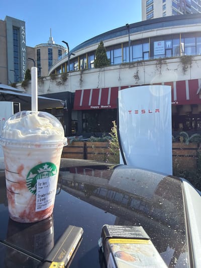 This is a vertical, outdoor, daytime photograph taken in Büyükçekmece, Turkey. The scene features a Tesla charging station prominently in the foreground, with a Starbucks drink to its right. Behind the charging station, a building with red and white striped awnings and large glass windows can be seen, labeled "Happy Moon's." Further in the background, modern buildings and a clear blue sky suggest a bright, sunny day.
On the Starbucks cup, text indicates "Strawberries&Crm Frap" and "p Grande," along with "Starbucks Coffee" and a time stamp of "18:00:31." A pack of cigarettes, featuring prominent health warnings in Turkish, sits to the right of the drink. Visible text on the building includes "KAYA MILLENI'" and a dental clinic's name and phone number. The overall impression is of a casual day, possibly late afternoon given the time stamp on the Starbucks cup, in a modern urban setting with commercial establishments. There is no visible activity or people in the image.