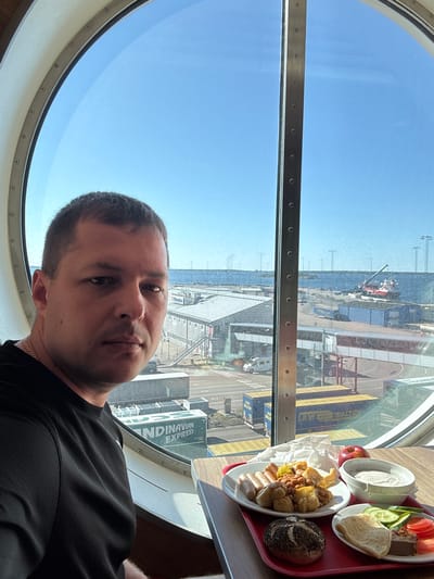 A man sits at a table with a meal in front of him, by a large, circular window. The man is wearing a black t-shirt and a silver chain. He is looking towards the camera. On the table, a red tray holds a plate with sausages, beans, and potatoes, a bread roll, a small plate with a bread roll, cucumber slices, tomato slices, and a piece of meat. There is also a red apple and a bowl of white yogurt. Through the window, a port is visible with industrial buildings, shipping containers, and a red and white ship in the water. The sky is clear and blue. This scene is likely in the city of Karlskrona, Sweden, as suggested by the "Scandinavian Express" shipping container, a known ferry operator.