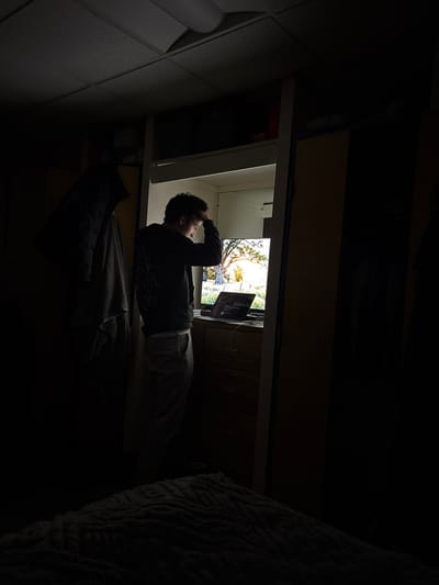 A dimly lit room contains a man lying down, looking at a computer screen. The screen displays an image of trees and sunlight. The man's hand is raised to his head, appearing thoughtful. A partially visible bed with a textured blanket is on the right, and dark furniture frames the top of the shot. A dark-colored jacket lies on the surface under the man. The overall scene has a quiet, contemplative mood.