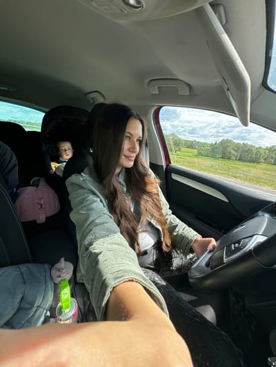 The interior of a car, presumably in motion, reveals a woman in the driver's seat, likely taking a selfie. She has long brown hair and is wearing a light green jacket. She is holding the steering wheel with her left hand. In the back seat, a baby in a car seat is visible, along with a pink bag, a water bottle, and a blue blanket. The car's interior is a mix of black and gray. Through the car window, a green field and some trees can be seen under a cloudy sky. The specific location within the city of Kremyonki, Russia, is not discernible.