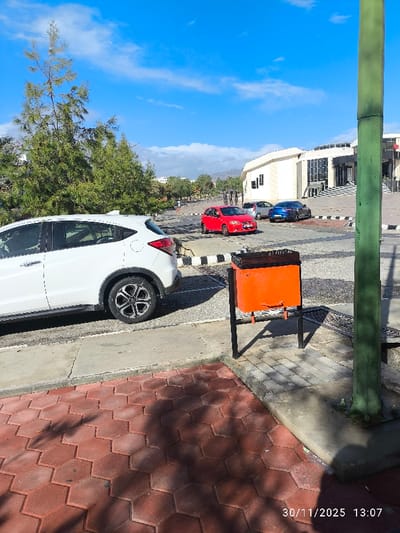 The image depicts an outdoor scene in Gönyeli, Northern Cyprus / Cyprus, captured on November 30, 2025, at 13:07. The foreground features a pedestrian area paved with red hexagonal interlocking pavers, partially shaded, adjacent to a concrete sidewalk. A white Honda HR-V SUV, with distinctive multi-spoke wheels, is parked facing left on the concrete. To its right, an orange, rectangular metal trash bin with a black liner is visible, mounted on a black metal frame. A vertical green pole occupies the far right of the frame.
Beyond the sidewalk, an asphalt parking area is present, separated by black and white striped curbstones. Three vehicles are parked in this area: a red Mitsubishi Lancer sedan, a silver sedan positioned behind the red car, and a blue sports car/coupe further back. In the midground, various green trees are visible, including tall, slender evergreens.
The background is dominated by a large, light-colored, modern-style building, likely institutional, featuring multiple windows and a prominent entrance with a wide set of steps. Distant hills or mountains are visible under a clear blue sky with scattered white clouds. No individuals are present in the frame. The scene is well-lit by direct sunlight, casting shadows towards the right and rear of the objects.