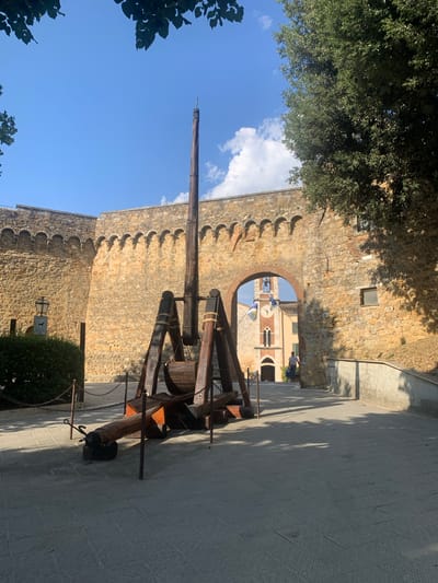 The image depicts a view through an arched stone gateway, likely part of a historic fortification, in San Quirico d'Orcia, Italy. In the foreground, a large wooden catapult-like structure is prominently displayed, suggesting it might be a historical exhibit or replica. The catapult has a long wooden arm, a heavy base, and is secured with chains.

Beyond the gateway, a glimpse of a town square is visible. A clock tower with flags and a bell is a central feature. A person in a blue shirt is standing on a pathway, looking towards the tower. The architecture visible through the archway suggests a traditional Italian town.

The scene is bathed in bright sunlight, casting shadows from trees onto the stone pavement. The sky is a clear blue with a few white clouds. The stone walls of the fortification are aged and made of rough-hewn stones, with crenellations along the top. The overall atmosphere is one of historical charm and a sunny day. The time of day appears to be late morning or early afternoon, given the strong sunlight and the angle of the shadows. There is a small sign with an icon of a dog on it near a bush.