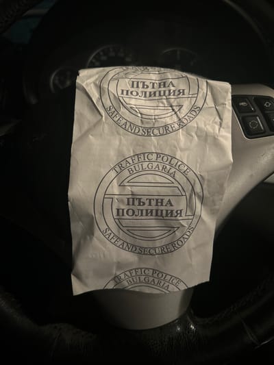 An overhead close-up shows a crumpled piece of white paper resting on the steering wheel of a car. The steering wheel is black, likely leather, and partially visible in the dim lighting of the car's interior. On the left side, a section of the car's dashboard with silver-colored control buttons (possibly for media or phone) is visible.
The paper features repeated circular stamps printed in dark blue or black ink. The text on these stamps is in both Bulgarian Cyrillic and English. Visible text includes "ПЪТНА ПОЛИЦИЯ" (Bulgarian for Traffic Police), "TRAFFIC POLICE", "BULGARIA", and "SAFE AND SECURE ROADS". The paper appears to be an official document or notice from the Bulgarian Traffic Police, possibly left on the vehicle.
The lighting in the image is low, suggesting it might be nighttime or an indoor setting with minimal illumination. The context provided indicates the location is Sofronievo, Bulgaria, which aligns with the text on the paper. No people are visible, and no specific activity beyond the placement of the paper is depicted.