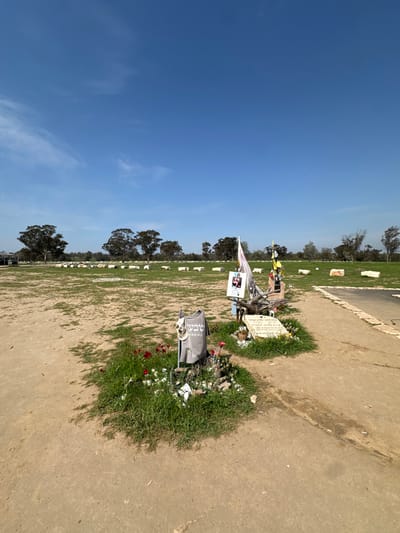 The image shows a memorial site in the city of Re'im, Israel, located at the coordinates 31.398 latitude and 34.471 longitude. The memorial is surrounded by dry, light brown soil and sparse patches of green grass. There are several trees in the background to the left of the frame. The memorial consists of a cluster of white stones, flowers, and a few items including a small flag, a photograph, and a t-shirt.  A white stone inscribed with Hebrew writing is surrounded by red flowers. The site is situated on an open, flat area in the city.  The sky is a bright blue with some wispy clouds in the distance.  The overall scene appears to be a somber and respectful memorial site.