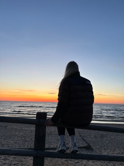 A woman in a dark jacket, dark leggings, and white shoes is standing on a wooden fence at the beach in Kalngale, Latvia. She is leaning forward and looking towards the beach. In the background is the sky and the sea at sunset, with a gradient of blue to orange. The beach sand is visible below the fence.
