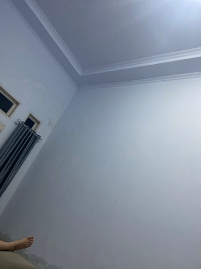 The image depicts an interior view of a room. The walls are painted a light color, with some water stains visible near the floor and ceiling. A portion of the ceiling with decorative molding is visible in the upper part of the image. On the left side, two windows are present. One window is partially visible at the top left, and another is larger, with dark grey curtains hanging in front of it. A person's foot is visible in the lower left corner of the image, resting on a surface that appears to be a bed or sofa. The scene suggests a residential setting. Given the location of Walennae, Indonesia, this scene is likely a bedroom or living area in a home within that region.