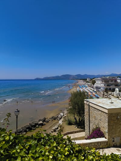 A high-angle, wide shot captures a bright, sunny day at a beach resort in Sperlonga, Italy. The azure sky dominates the upper portion of the image, meeting the deep blue of the Tyrrhenian Sea at the horizon. The sea is calm, with gentle waves lapping at a sandy beach.

The beach is lined with a dense array of colorful umbrellas and lounge chairs, suggesting a popular tourist destination. A few people can be seen walking along the shoreline and in the shallow water. Along the edge of the beach, a rocky embankment separates the sand from a grassy area with trees and a few buildings. One prominent building, identified as the "AURORA HOTEL SPERLONGA," is white with multiple levels and balconies.

In the foreground, lush green ivy covers a stone wall and railing, framing the view of the beach and sea. A vintage-style street lamp stands on the embankment near the water. The overall scene depicts a vibrant coastal vacation spot.